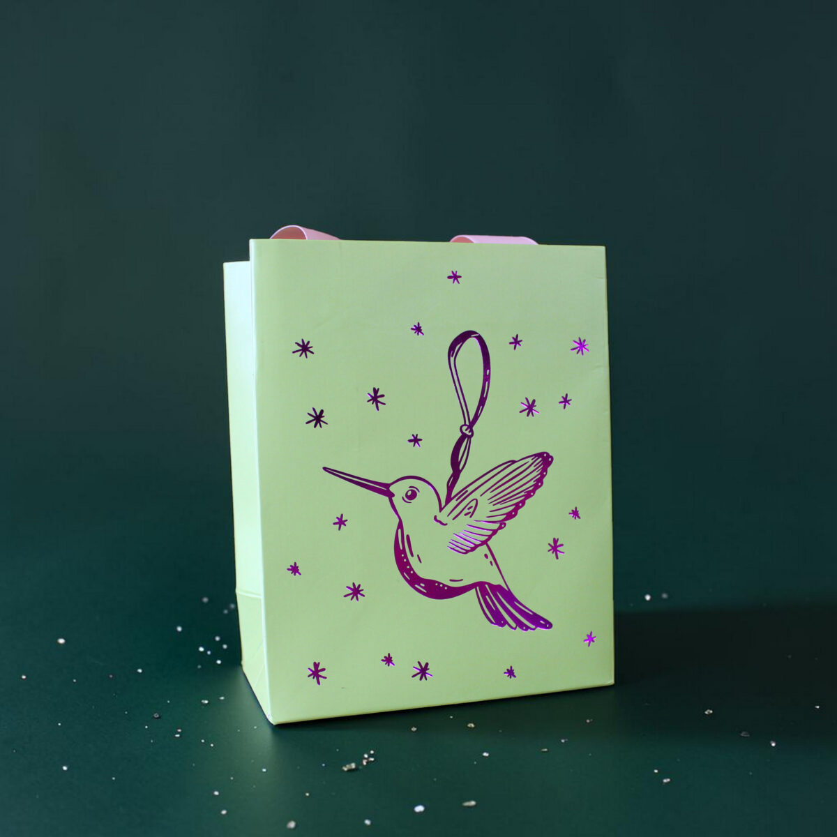 4179geschenktaschesvogel02 Avayves Feine Papeterie Und Geschenke Für Babys Und Kids Pale green gift bag with a purple bird illustration and star shapes on a dark teal background, with white confetti around it.