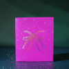 4180geschenktaschemschleife02 Avayves Feine Papeterie Und Geschenke Für Babys Und Kids Pink gift box with a glitter bow sits on a dark teal surface, sprinkled with confetti scattered around.