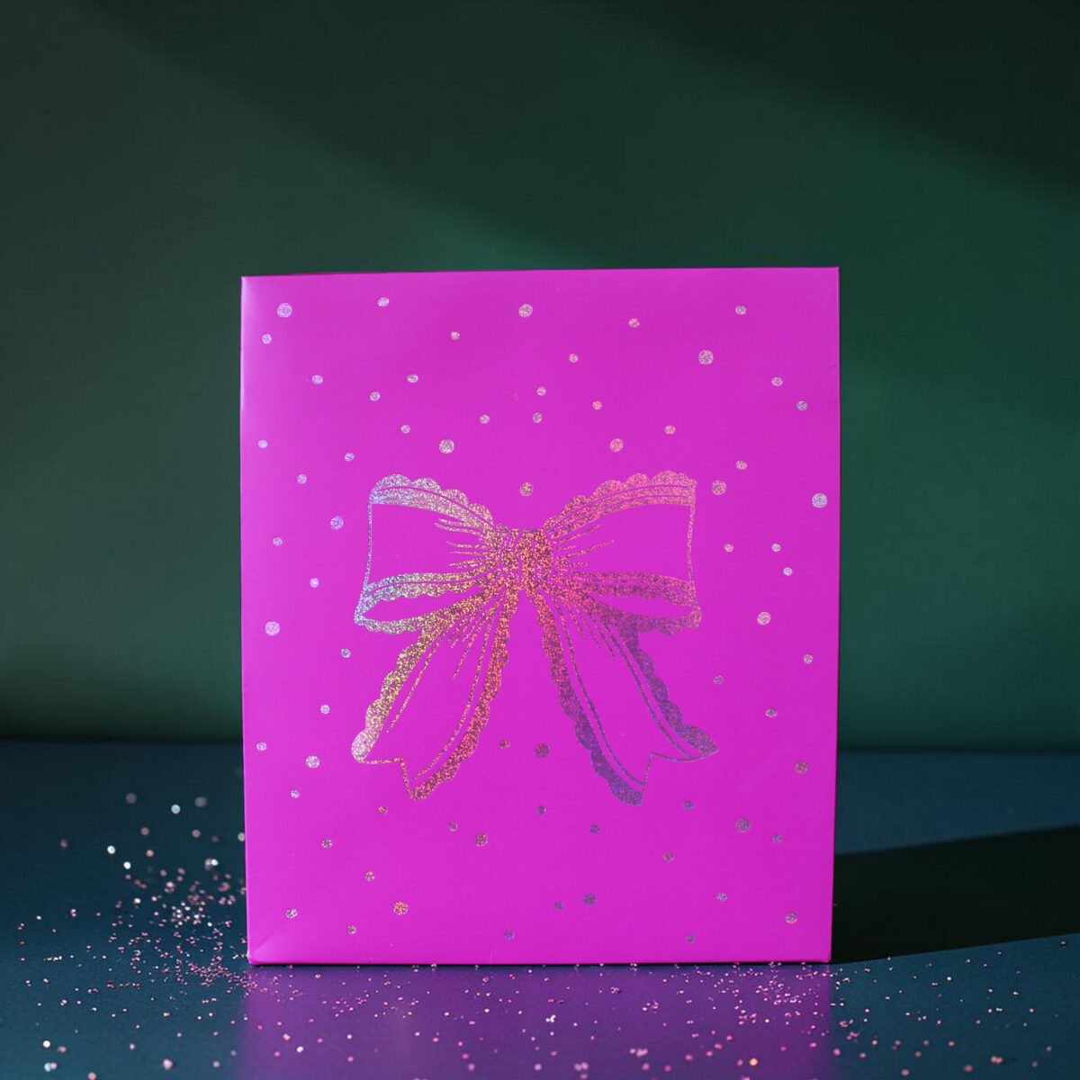 4180geschenktaschemschleife02 Avayves Feine Papeterie Und Geschenke Für Babys Und Kids Pink gift box with a glitter bow sits on a dark teal surface, sprinkled with confetti scattered around.