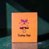 4181geschenktaschempilz02 Avayves Feine Papeterie Und Geschenke Für Babys Und Kids Orange gift box with a purple bell illustration and 'Frohes Fest' text on a glittery green surface, festive mood.
