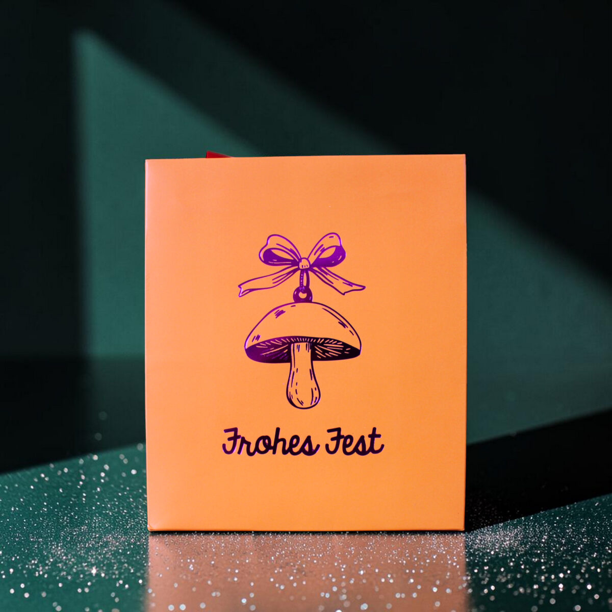 4181geschenktaschempilz02 Avayves Feine Papeterie Und Geschenke Für Babys Und Kids Orange gift box with a purple bell illustration and 'Frohes Fest' text on a glittery green surface, festive mood.