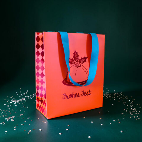 Pink gift bag with bright blue handles on a dark green surface, scattered white confetti around it.