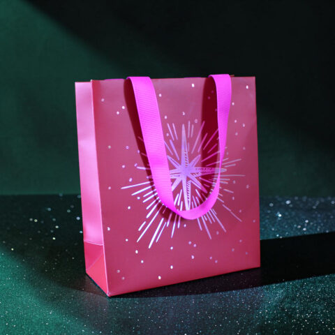 Pink gift bag with bright pink handles on a dark glittery background, featuring white starburst pattern.