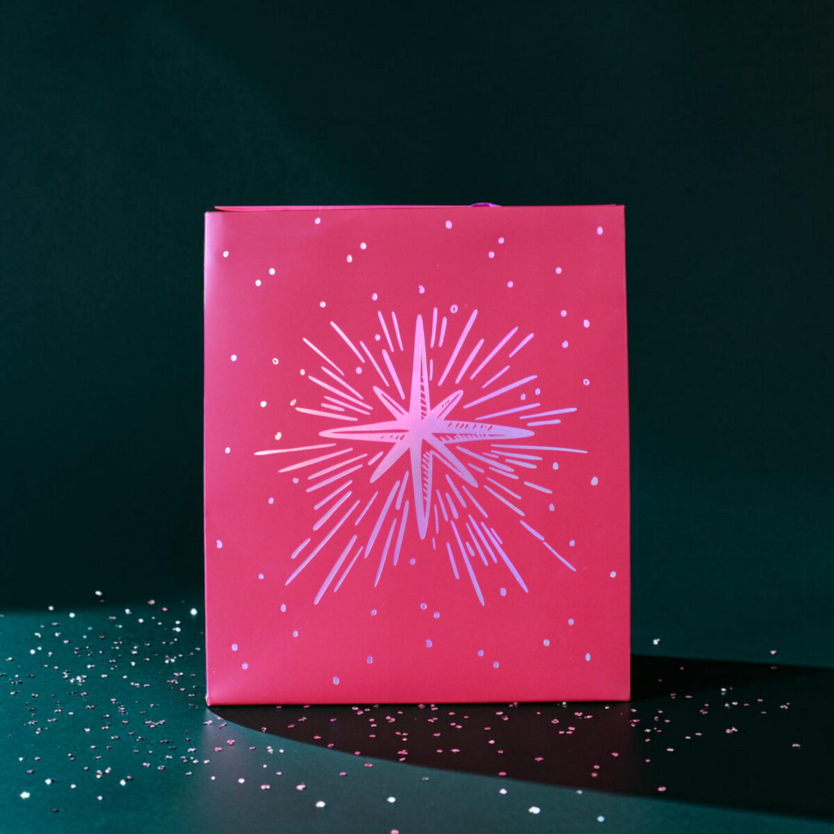 4183geschenktaschemstern02 Avayves Feine Papeterie Und Geschenke Für Babys Und Kids Pink gift box with white firework-like pattern on a dark teal background, surrounded by scattered confetti.