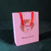 Pink gift bag with red handles against a dark speckled background, showing a festive design and partial 'Merry Christmas' text on the bag.