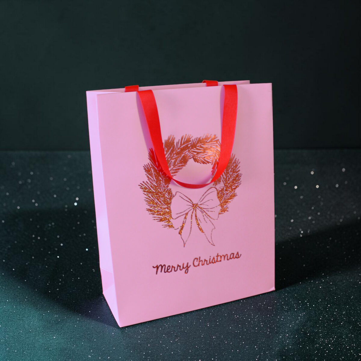 4184geschenktaschelkranz Avayves Feine Papeterie Und Geschenke Für Babys Und Kids Pink gift bag with red handles against a dark speckled background, showing a festive design and partial 'Merry Christmas' text on the bag.