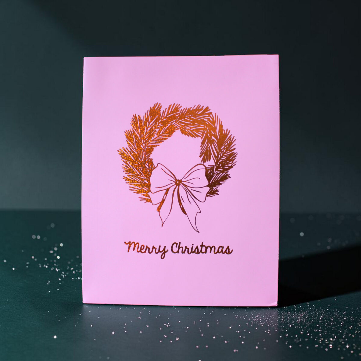 4184geschenktaschelkranz02 Avayves Feine Papeterie Und Geschenke Für Babys Und Kids Pink Christmas card with a pine wreath and bow, reading 'Merry Christmas', set on a dark green background with confetti specks.