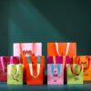 Allegeschenktaschenxmas26 Avayves Feine Papeterie Und Geschenke Für Babys Und Kids Colorful gift bags with bright ribbons lined up at the bottom of a dark green background.