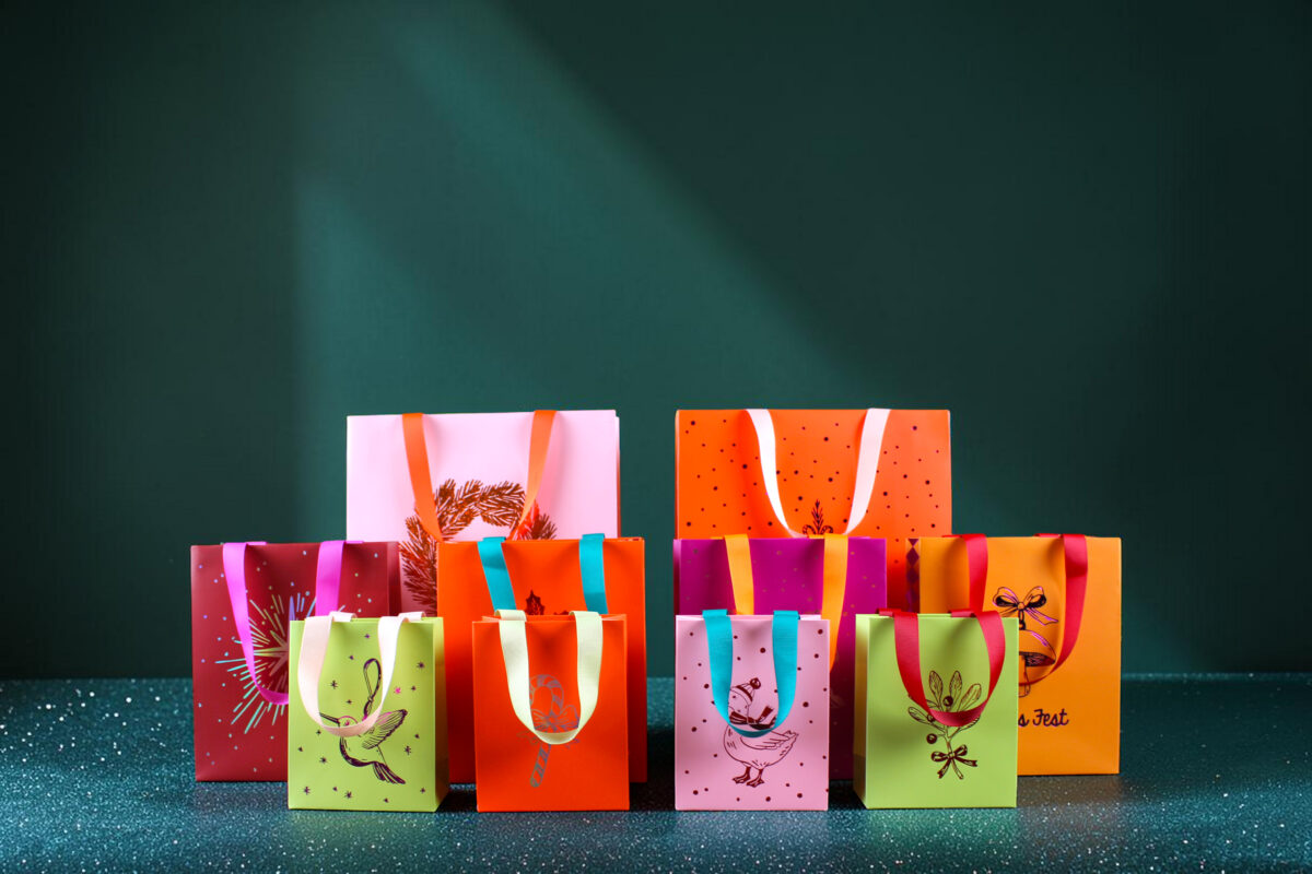 Allegeschenktaschenxmas26 Avayves Feine Papeterie Und Geschenke Für Babys Und Kids Row of brightly colored gift bags with ribbons lined up on a speckled floor in front of a dark green wall.