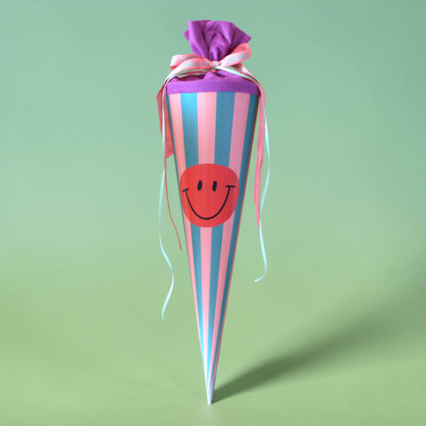 Tall striped party cone in pink and blue with an orange smiley face sticker and pastel ribbons at the top, set against a mint-green background.