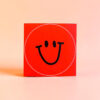 5899aufklebersmileneonrot Avayves Feine Papeterie Und Geschenke Für Babys Und Kids Red square box featuring a bold black smiley face inside a white circular outline on a bright red background.