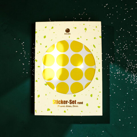 Cream-colored packaging with a circular window revealing yellow polka-dot stickers; green confetti pattern on the box edge.