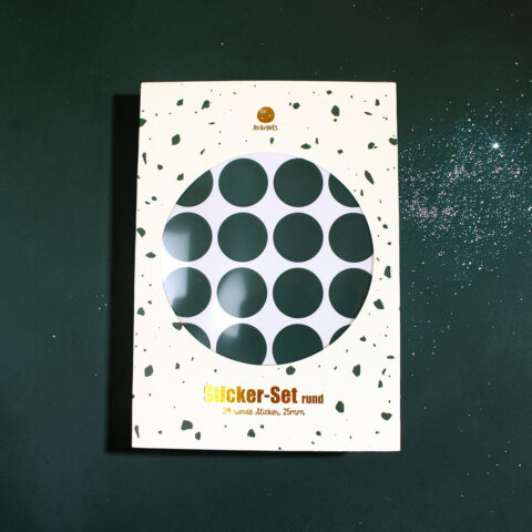 White box packaging for a Sticker-Set, featuring a circular window showing dark green polka-dotted sheet inside and scattered green confetti design on the box edges.