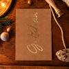 Brown holiday greeting card with gold 'Christmas' script on a wooden table, surrounded by pine, candle, rope, and natural decorations.
