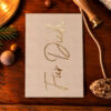 1203pkfuerdichgoldfolie Avayves Feine Papeterie Und Geschenke Für Babys Und Kids Beige greeting card with gold cursive text on a wooden table, surrounded by holiday decorations like pine branch, nuts, rope, and a stamping tool/mini brass press beside it.