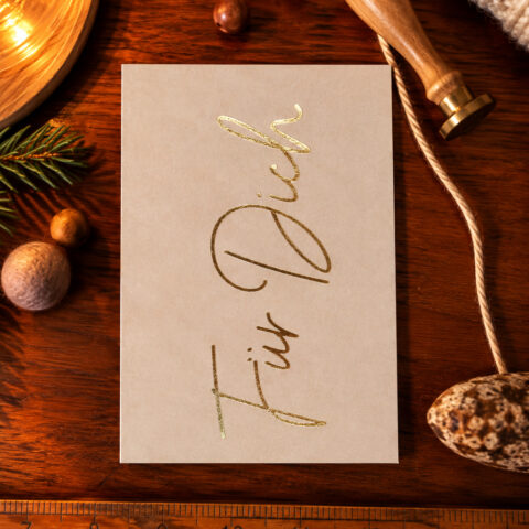 Beige greeting card with gold cursive text on a wooden table, surrounded by holiday decorations like pine branch, nuts, rope, and a stamping tool/mini brass press beside it.