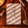 1211pkhohohostreifenrosarost Avayves Feine Papeterie Und Geschenke Für Babys Und Kids Gold glittery cursive 'Merry' on a pink-beige diagonal-striped card placed on a wooden surface, surrounded by holiday props (pine branch, cinnamon, rope).