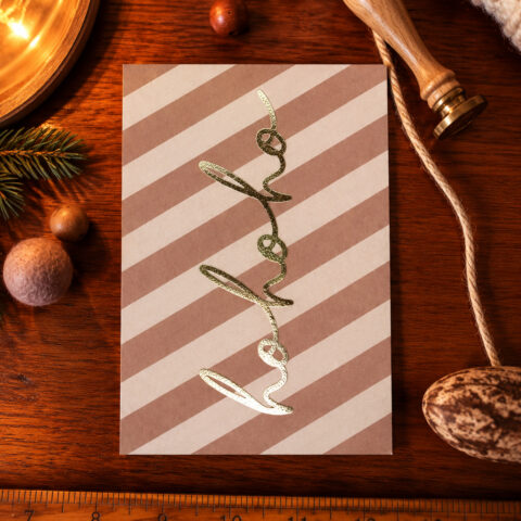 Gold glittery cursive 'Merry' on a pink-beige diagonal-striped card placed on a wooden surface, surrounded by holiday props (pine branch, cinnamon, rope).