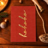 Red Christmas card with gold cursive text on a wooden table beside holiday decorations.