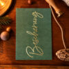 Green greeting card with gold cursive text 'Greetings' on a wooden table, surrounded by holiday decor like pine, rope, and ornaments.