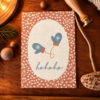 Christmas card on a wooden surface with a white oval center showing blue mitten illustrations and 'hohoho' in blue cursive, brown polka-dot background.