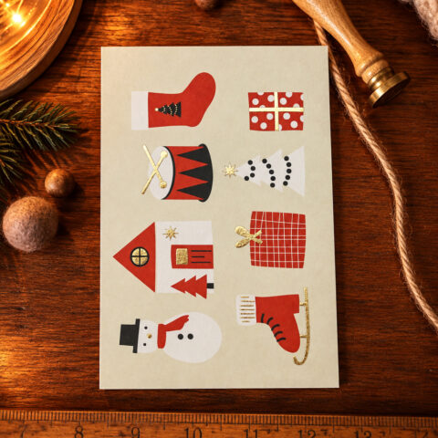 Beige holiday card on a wooden surface showing paper cutouts: red mitten, polka-dot gift, drum with sticks, white Christmas tree, red house, plaid gift bag, snowman, red ice skate.