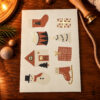Seasonal card on a wooden table featuring stylized icons: stocking, gifts, drum, tree, house, snowman, and ice skate.
