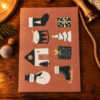 1256pktrommelhausstrumpfdunkelrosagruengold Avayves Feine Papeterie Und Geschenke Für Babys Und Kids Festive collage on a brown card showing cutout shapes: sock, present, drum, stacked tree, house, plaid gift bag, snowman, Christmas tree slices, and ice skate.