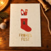 White Christmas card featuring a red stocking with Santa and gifts peeking out, gold confetti, and the gold caption 'FROHES FEST' on a wooden table. A pine branch and twine are visible nearby.