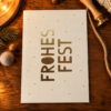 1258pkfrohesfest Avayves Feine Papeterie Und Geschenke Für Babys Und Kids Cream holiday card with gold 'FROHES FEST' text and small red dots on a wooden table amid pine and twine, festive scene.
