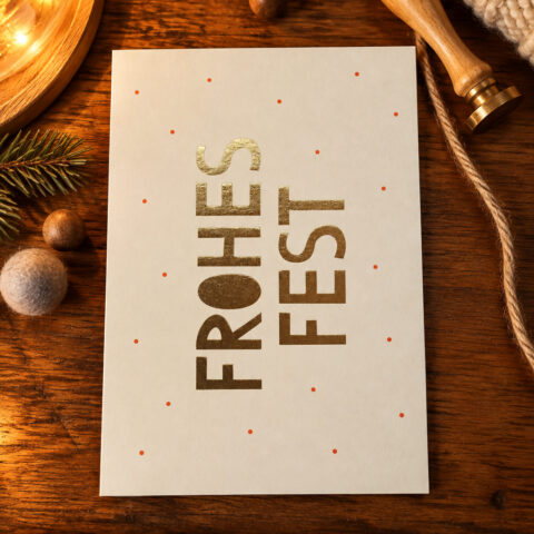 Cream holiday card with gold 'FROHES FEST' text and small red dots on a wooden table amid pine and twine, festive scene.