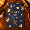 Blue greeting card with gold stars and the text 'FROHES FEST' on a wooden table, evokes festive holiday wishes.