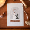1275pkfrohesfestschneekugel Avayves Feine Papeterie Und Geschenke Für Babys Und Kids Flat lay of a holiday card with a snowman and trees on a wooden surface, tied with twine; measuring tape at the bottom hints at size.