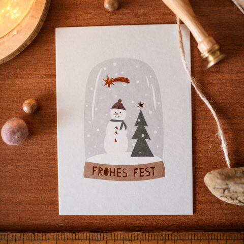 Flat lay of a holiday card with a snowman and trees on a wooden surface, tied with twine; measuring tape at the bottom hints at size.