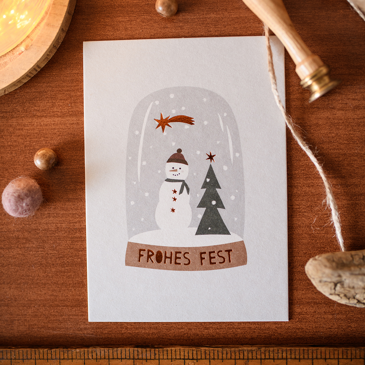 Snow globe-style card with a snowman and a Christmas tree inside a dome, banner reads 'FROHES FEST' on a wooden desk surface