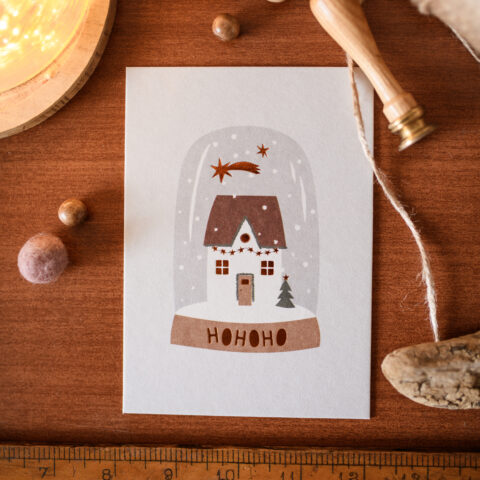 Close-up of a holiday card showing a snowy house illustration, with 'HOHOHO' at the bottom, on a wooden desk beside a measuring ruler and twine.
