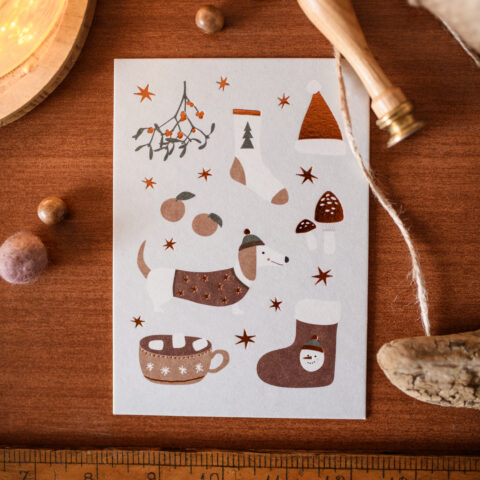 Flat-lay holiday card featuring illustrated mug, snowman, tree and stars on a wooden table with a measuring ruler along the bottom edge.