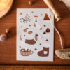 Whimsical holiday card on a wooden surface showing a dog, stocking, mug, mushrooms, berries, a pine tree motif, and stars.