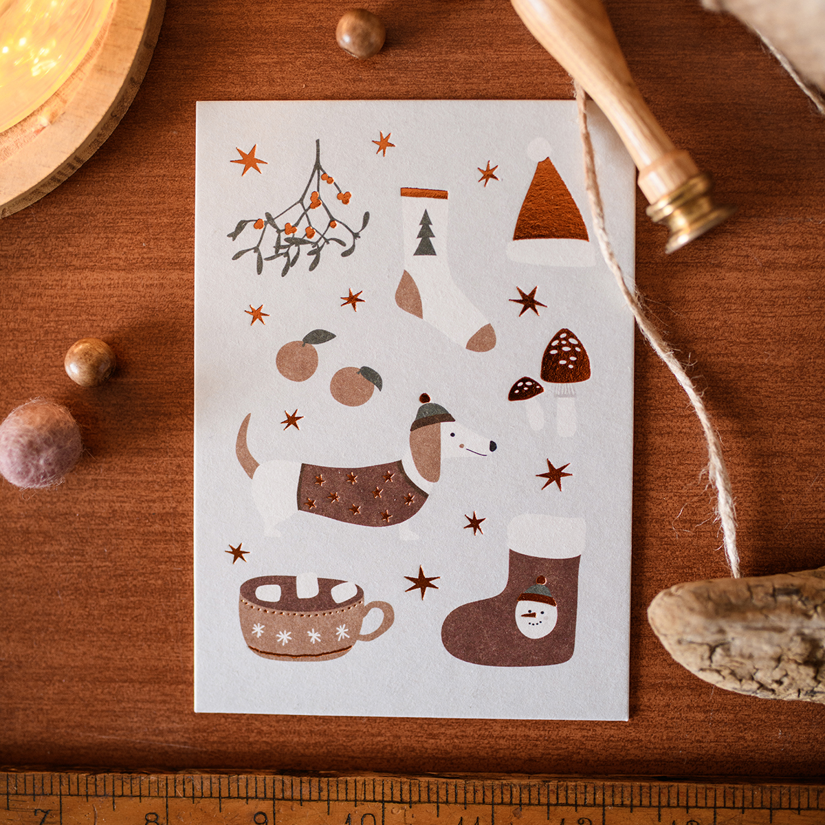 Whimsical holiday card on a wooden surface showing a dog, stocking, mug, mushrooms, berries, a pine tree motif, and stars.