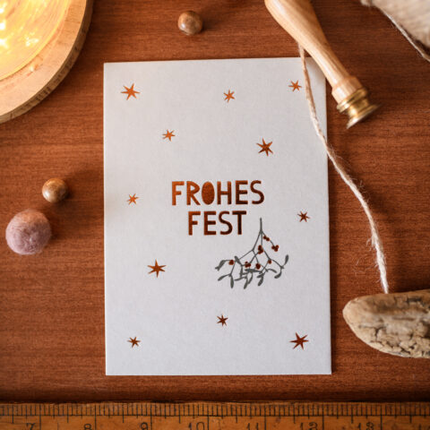 Overhead view of a white holiday card with copper stars on a wooden surface, with a string, a ruler at the bottom, and warm light nearby.