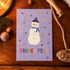 Purple holiday card with a snowman and 'FROHES FEST' text on a wooden table, surrounded by festive props (pine, ribbon, ruler).