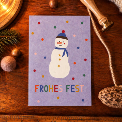 Purple holiday card with a snowman and 'FROHES FEST' text on a wooden table, surrounded by festive props (pine, ribbon, ruler).