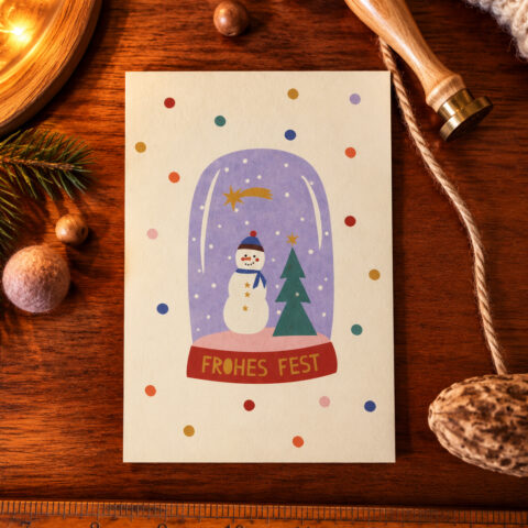 Colorful Christmas card with a snow globe scene showing a snowman and a Christmas tree, and the 'FROHES FEST' banner on a wooden desk.
