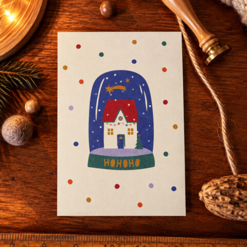 Seasonal card featuring a snow globe with a cozy house inside and 'HOHOHO' on the base, surrounded by colorful dots and a wooden background.