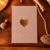 Beige greeting card with a gold heart centered, laid on a wooden desk with holiday props like pine needles and walnuts nearby.
