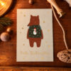 Christmas card with a brown bear in a green sweater featuring a snowman, surrounded by gold stars and 'Frohe Weihnachten' text on a wooden table surface.