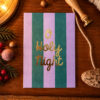 1365holynight Avayves Feine Papeterie Und Geschenke Für Babys Und Kids Card reading 'O Holy Night' in gold script on purple and green vertical stripes, placed on a wooden table with Christmas decorations nearby.