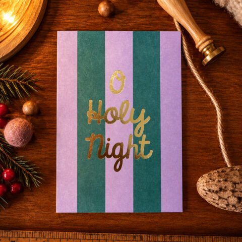 Card reading 'O Holy Night' in gold script on purple and green vertical stripes, placed on a wooden table with Christmas decorations nearby.