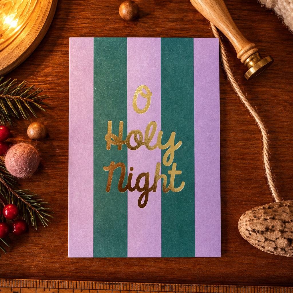 1365holynight Avayves Feine Papeterie Und Geschenke Für Babys Und Kids Card reading 'O Holy Night' in gold script on purple and green vertical stripes, placed on a wooden table with Christmas decorations nearby.