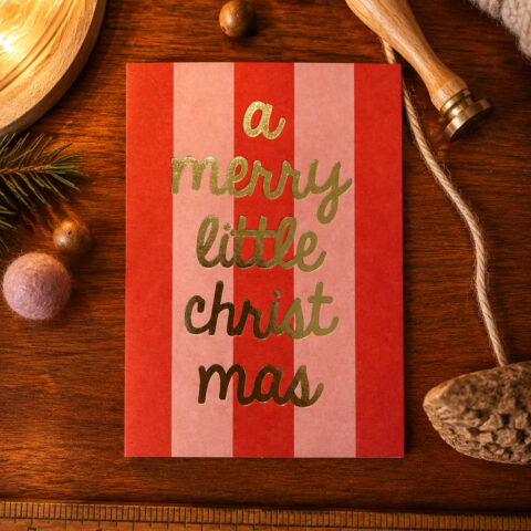 Card with gold script reading 'a merry little christmas' on red and pink striped background on a wooden table with pine cone and twine nearby.